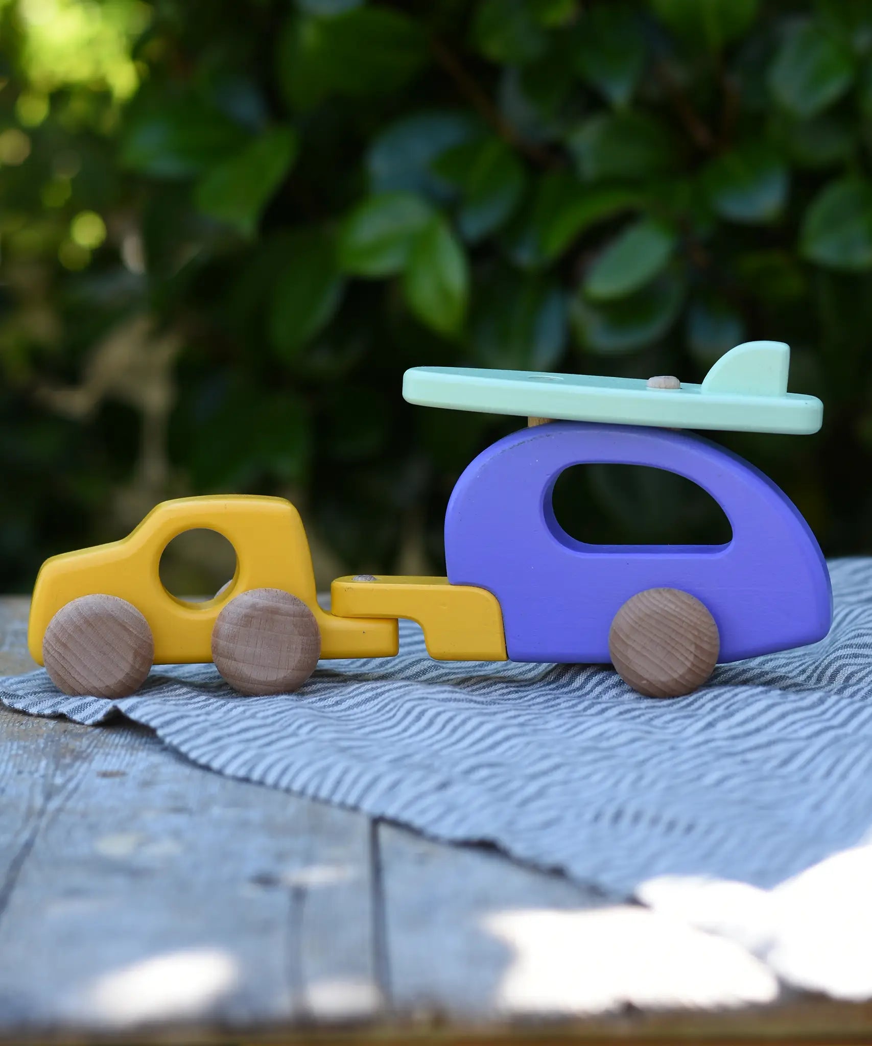 Bajo yellow car and purple camper wooden toy vehicle with green surfboard placed on a wooden surface outdoors.