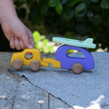 Child's hand pulling the Bajo yellow car and purple camper wooden toy vehicle with green surfboard along. .