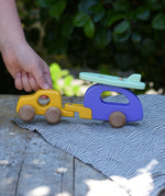 Child's hand pulling the Bajo yellow car and purple camper wooden toy vehicle with green surfboard along. .