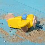 A yellow coloured Bajo toy plane with pilotplaced on a blue painted wooden surface covered by a scattering of sand. This plane is part of a wide range of Bajo wooden toy vehicles available at Babipur.