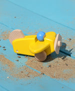 A yellow coloured Bajo toy plane with pilotplaced on a blue painted wooden surface covered by a scattering of sand. This plane is part of a wide range of Bajo wooden toy vehicles available at Babipur.