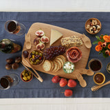A large Bambu bamboo serving and cutting board on a blue table cloth with various foot items on top for scale next to the bamboo spork honey dipper and tongs 