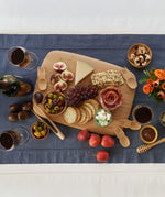 A large Bambu bamboo serving and cutting board on a blue table cloth with various foot items on top for scale next to the bamboo spork honey dipper and tongs 