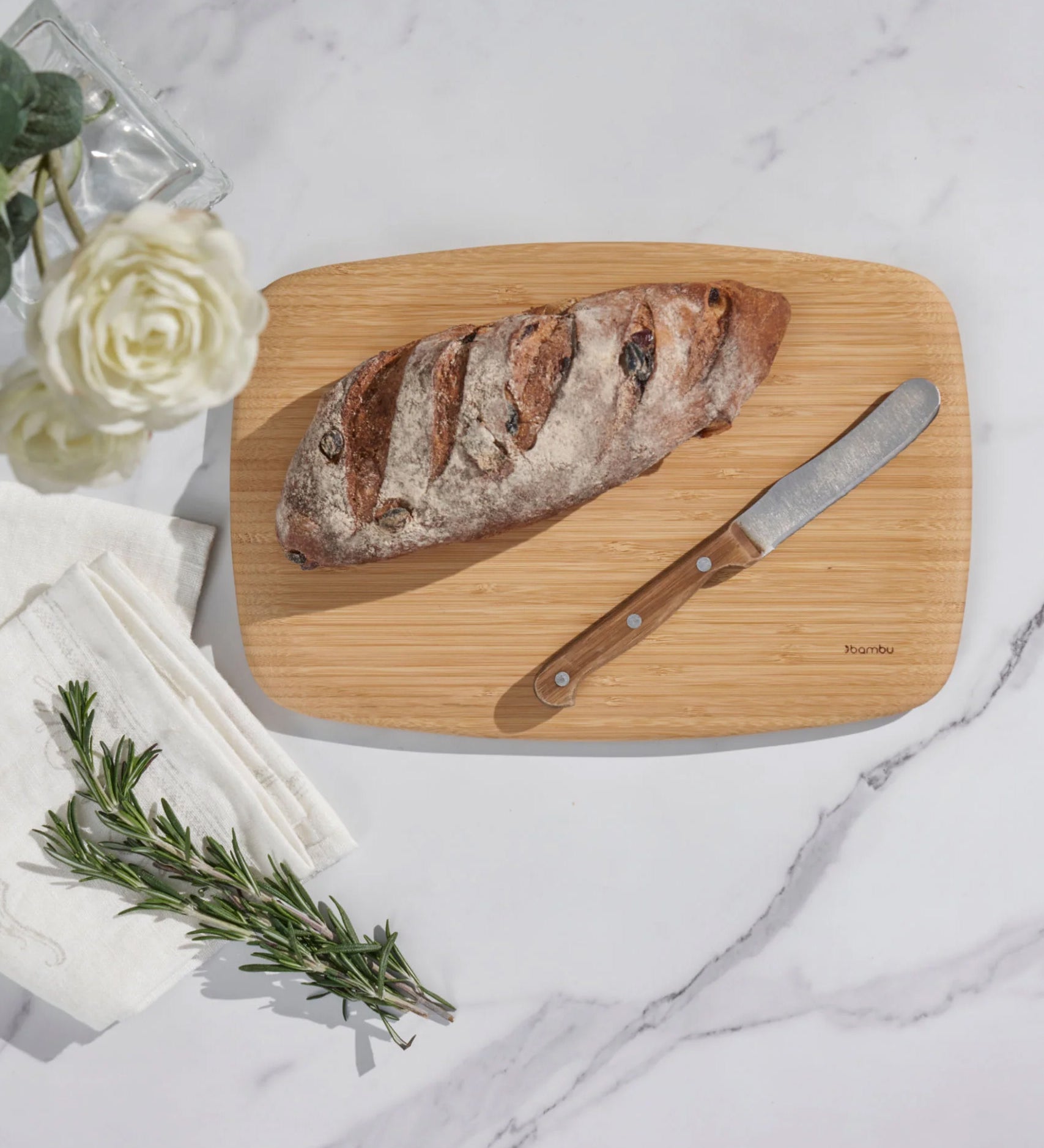 A Bambu medium serving and cutting bamboo board with a bread knife and a loaf of bread to show scale with a sprig of rosemary next to the board on a marble surface