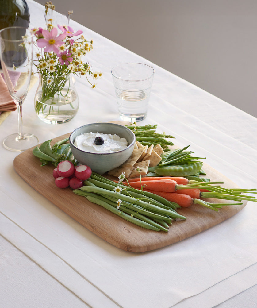 Close up view of the large Bumbu bamboo serving and cutting board on a white table cloth with various foods on top and showing the bamboo detail