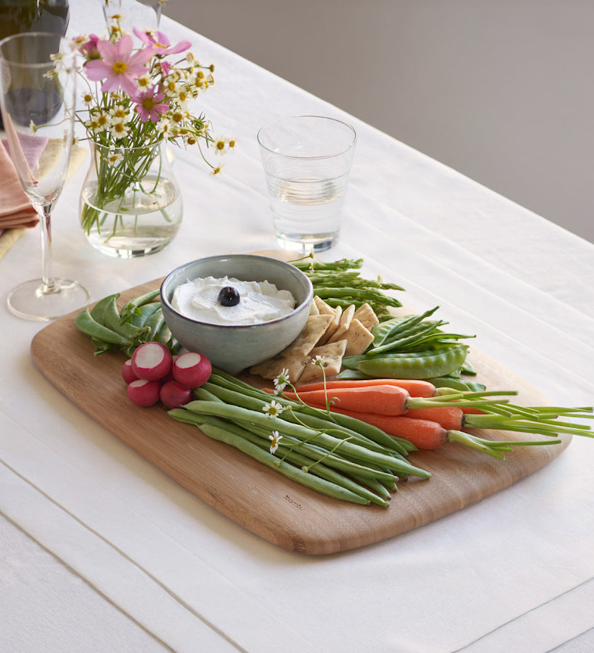 Close up view of the large Bumbu bamboo serving and cutting board on a white table cloth with various foods on top and showing the bamboo detail