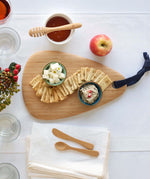 A Bambu Droplet shaped Artisan bamboo Board placed on a table on a white tablecloth. The boards has crackers and two ceramic bowls on it filled with cheese and humus. 