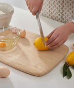 Adult hands cutting up a lemon on the large Bambu bamboo cutting board with a cracked egg in a small glass bowl on top showing the scale. 