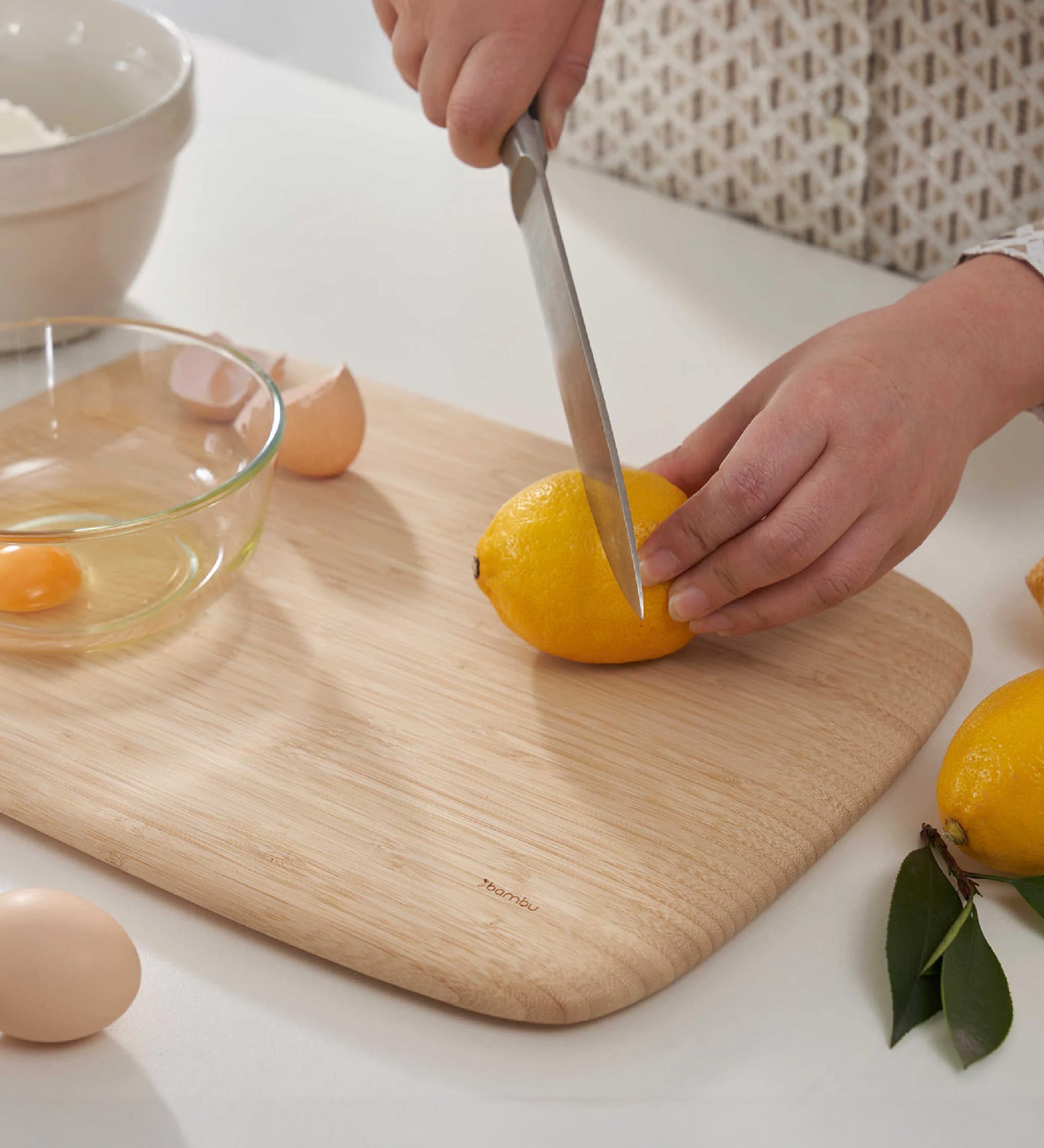 Adult hands cutting up a lemon on the large Bambu bamboo cutting board with a cracked egg in a small glass bowl on top showing the scale. 
