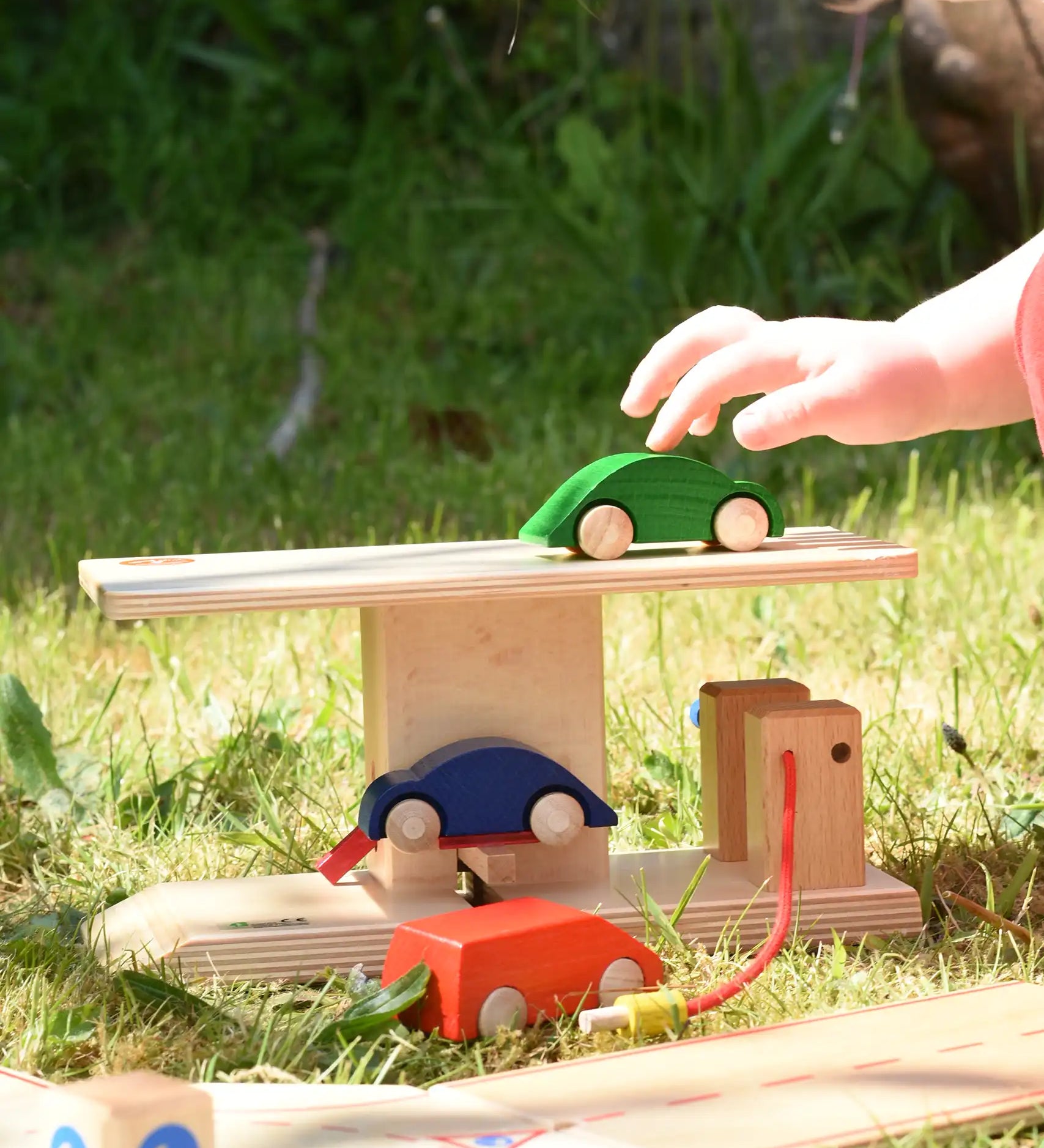 Beck wooden garage & petrol station for toy cars placed on grass outdoors. Child placing wooden car on the roof.
