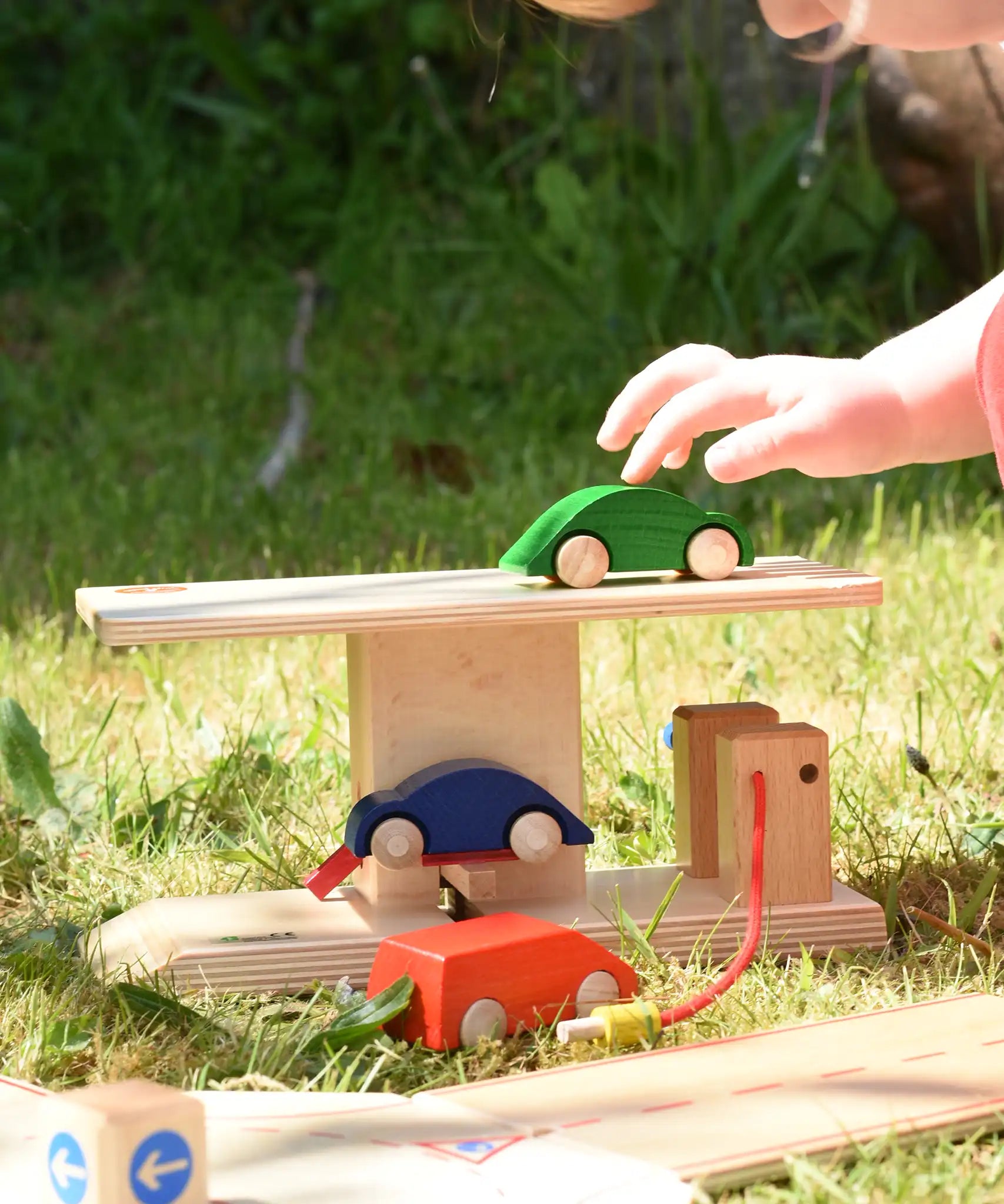 Beck wooden garage & petrol station for toy cars placed on grass outdoors. Child placing wooden car on the roof.