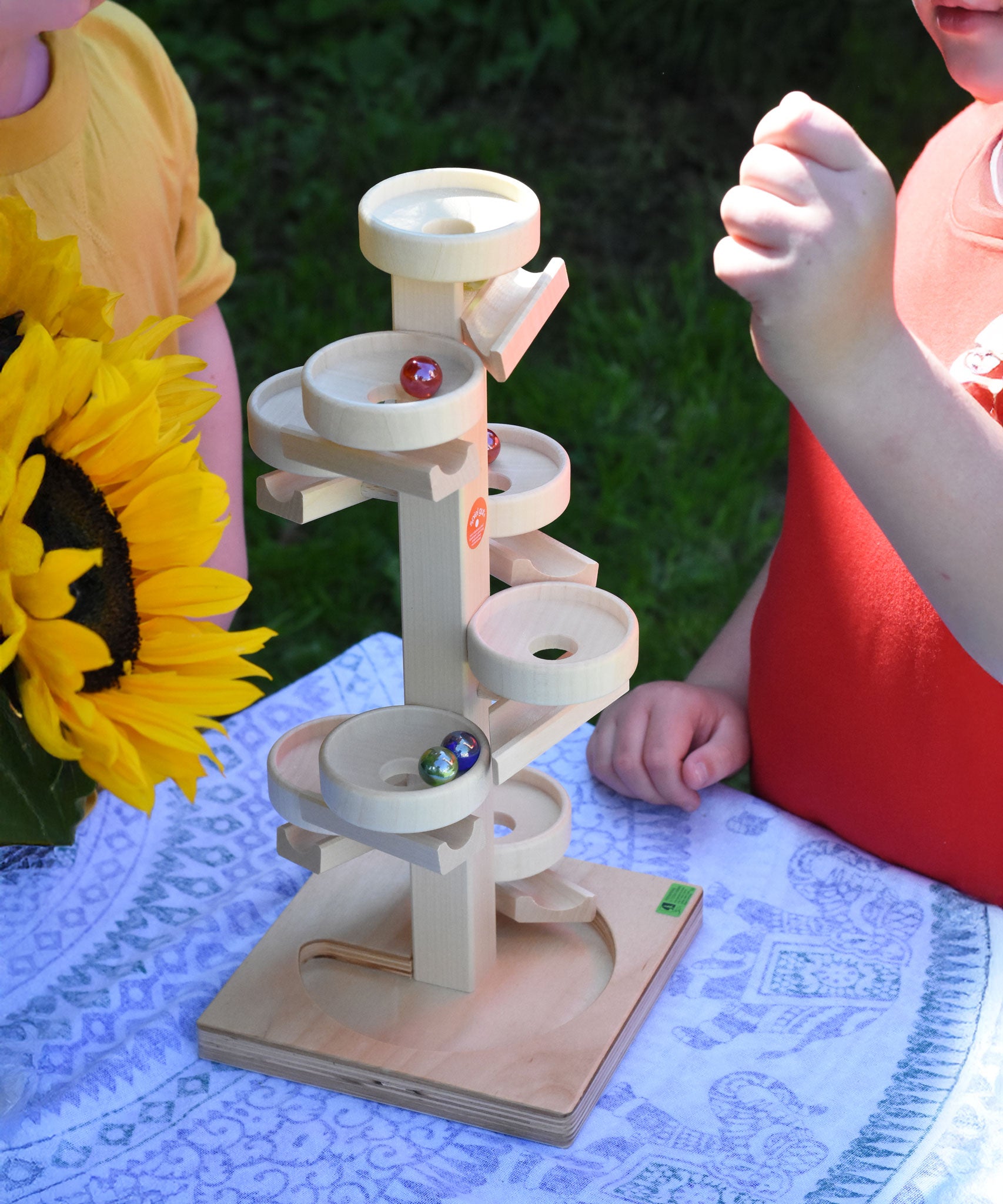 Two children playing with the Beck Marble Run Plate Tower which has been placed on a small table outdoors. A bunch of sunflowers can be seen to the side. 