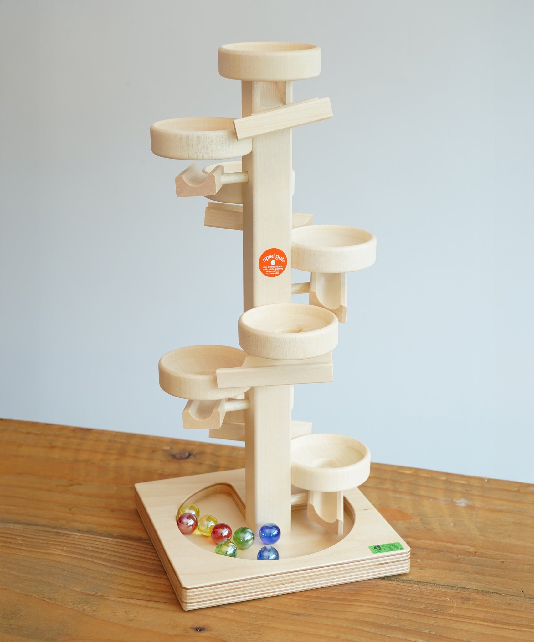 Beck Toys Maple Wood Marble Run Plate Tower placed on a wooden surface. The set comes with 8 coloured glass marbles that can be seen on the base. 