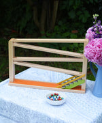 A Christof Beck Spielwaren Toys Chimes Marble Run Track placed on a small table outdoors. The coloured clay marbles have been placed in a small white ceramic bowl at the front. 