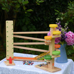 A collection of Beck Spielwaren wooden toys placed on a small table outdoors. The Wobbler tumbling toy, the run plate chimes marble run tower and the chimes marble run track. 