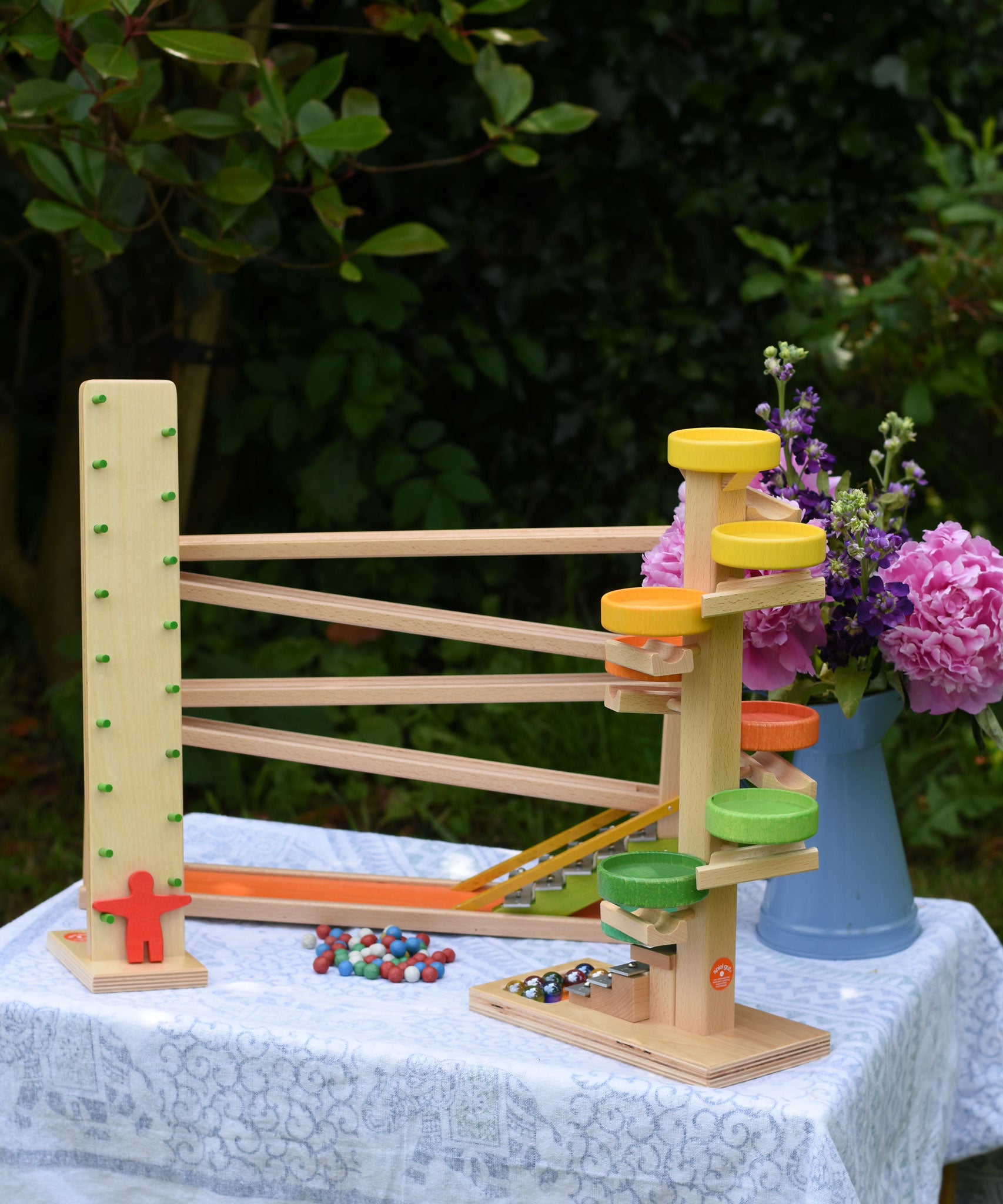 A collection of Beck Spielwaren wooden toys placed on a small table outdoors. The Wobbler tumbling toy, the run plate chimes marble run tower and the chimes marble run track. 