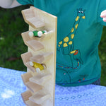 A close up of the Beck Toys Beechwood Cascading ‘Tower Running Track’ with Race Cars placed on a small table outdoors. Two wooden race cars can be seen zooming down the wooden track whilst a child is watching on. 