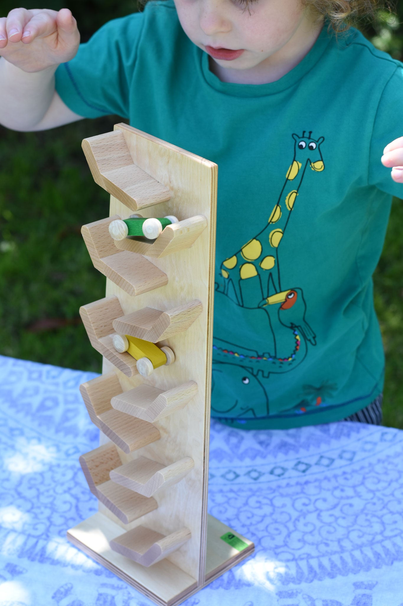 A close up of the Beck Toys Beechwood Cascading ‘Tower Running Track’ with Race Cars placed on a small table outdoors. Two wooden race cars can be seen zooming down the wooden track whilst a child is watching on. 
