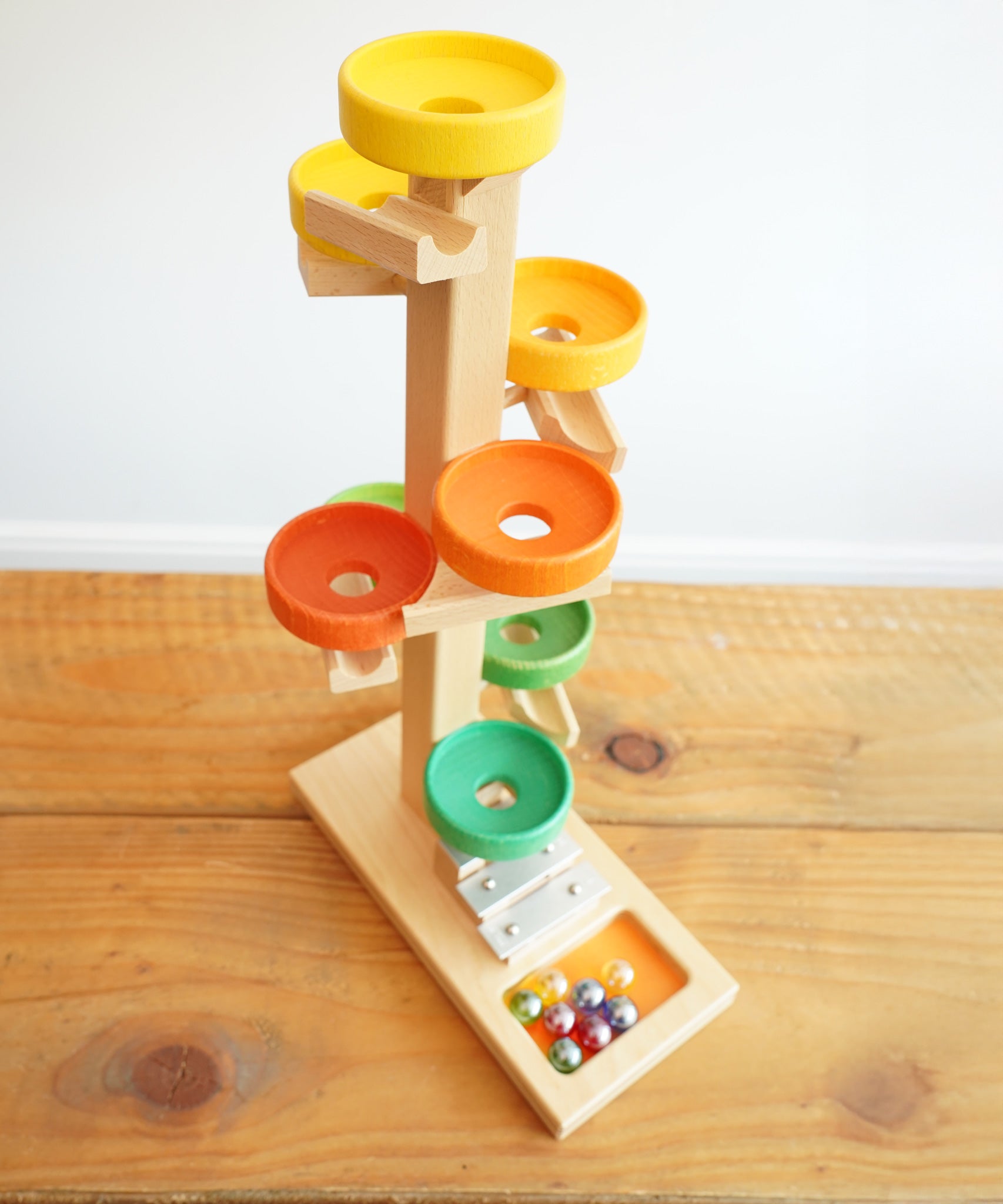 The Beck Toys Beech Wood Marble Run Plate Tower with Glockenspiel placed on a wooden surface. The set comes with 8 coloured glass marbles. 