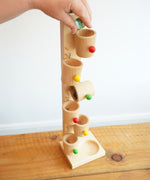 An adult's hand placing marbles into the first cup on the Beck Toys Beech Wood Roller Cup Marble Run tower. 