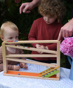 Two children playing with the Beck Toys Chimes Marble Run Track. An adult's hand can be seen placing some clay marbles on the track. 