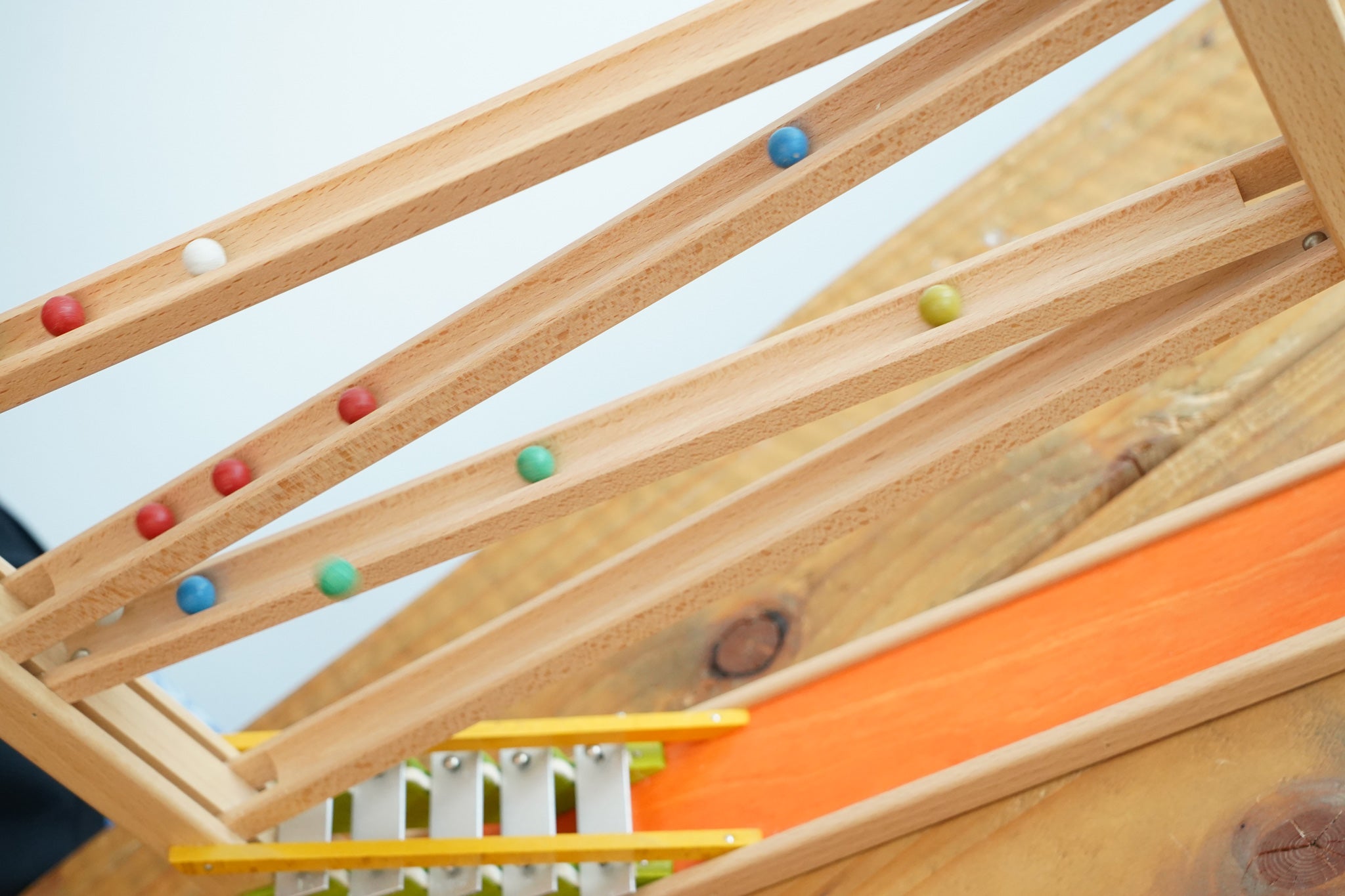 A side view of the Beck Toys Chimes Marble Run Track. Small colourful clay marbles can be seen blurred in motion, cascading down the wooden ramps. 