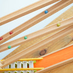 A side view of the Beck Toys Chimes Marble Run Track. Small colourful clay marbles can be seen blurred in motion, cascading down the wooden ramps. 