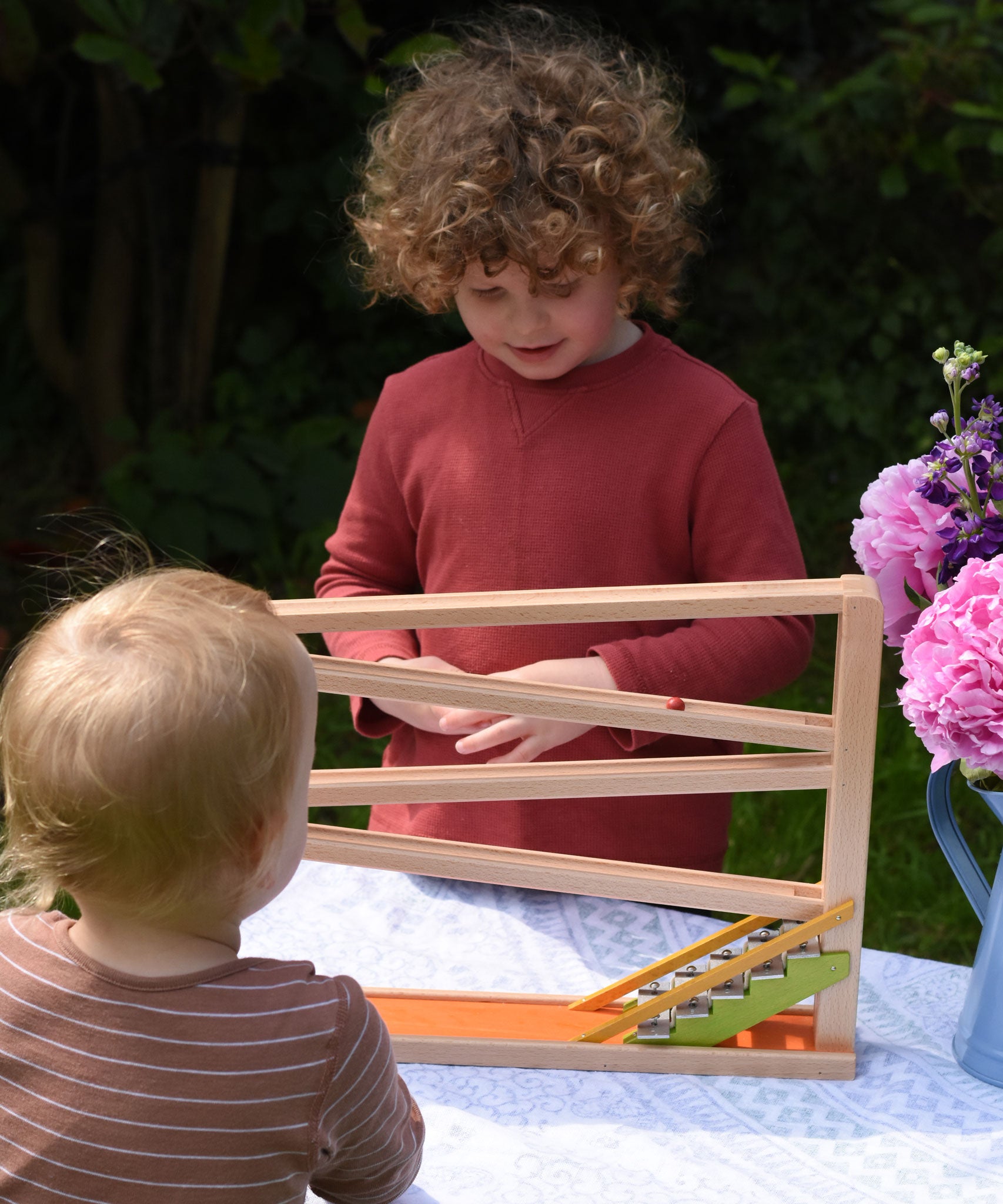 A child playing with the Beck Toys Chimes Marble Run Track. A younger child is standing in front observing one of the clay marbles going down the track.