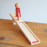 Beck Toys Nurmi Ramp Walker Wooden Toy placed on a wooden surface. The red colured wooden figure has been placed onto the ramp.