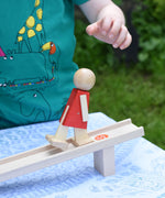 A close up of the Beck Toys Nurmi Ramp Walker Wooden Toy being played with outdoors. A child's hand can be seen just above the wooden walking figure that has been just placed on the wooden ramp. 