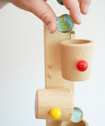 A close up of an adult's hand placing a glass marble into the first cup on the Beck Toys Beech Wood Roller Cup Marble Run. 