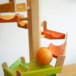 A orange coloured ball cascading down the track of the Beck Wooden musical chimes Ball Run. The ball is blurred in motion as it races down the wooden ramp pieces.