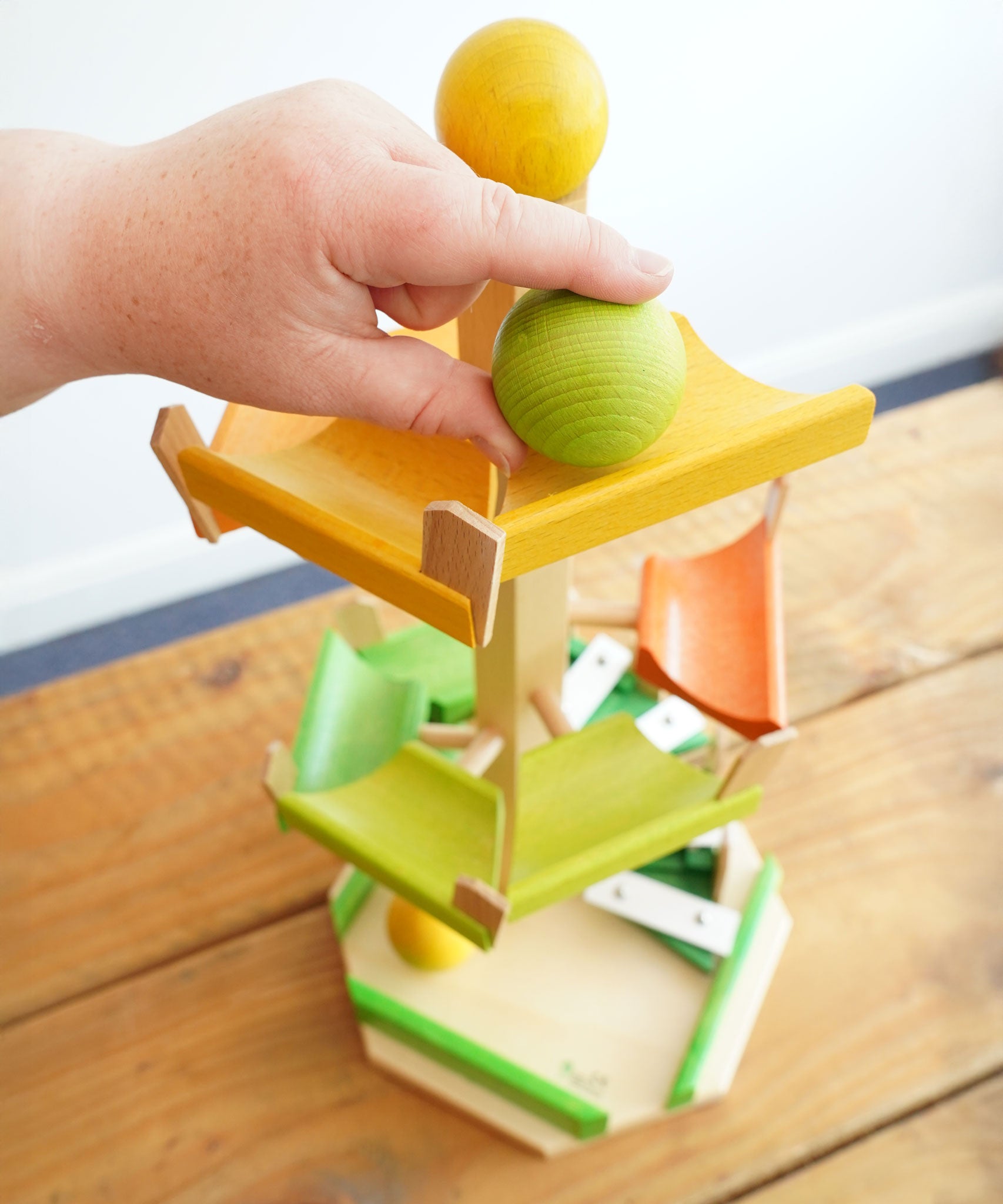 An adult's hand placing a green coloured ball on the track of the Beck Wooden musical chimes step tower ball run toy from Beck Toys. 