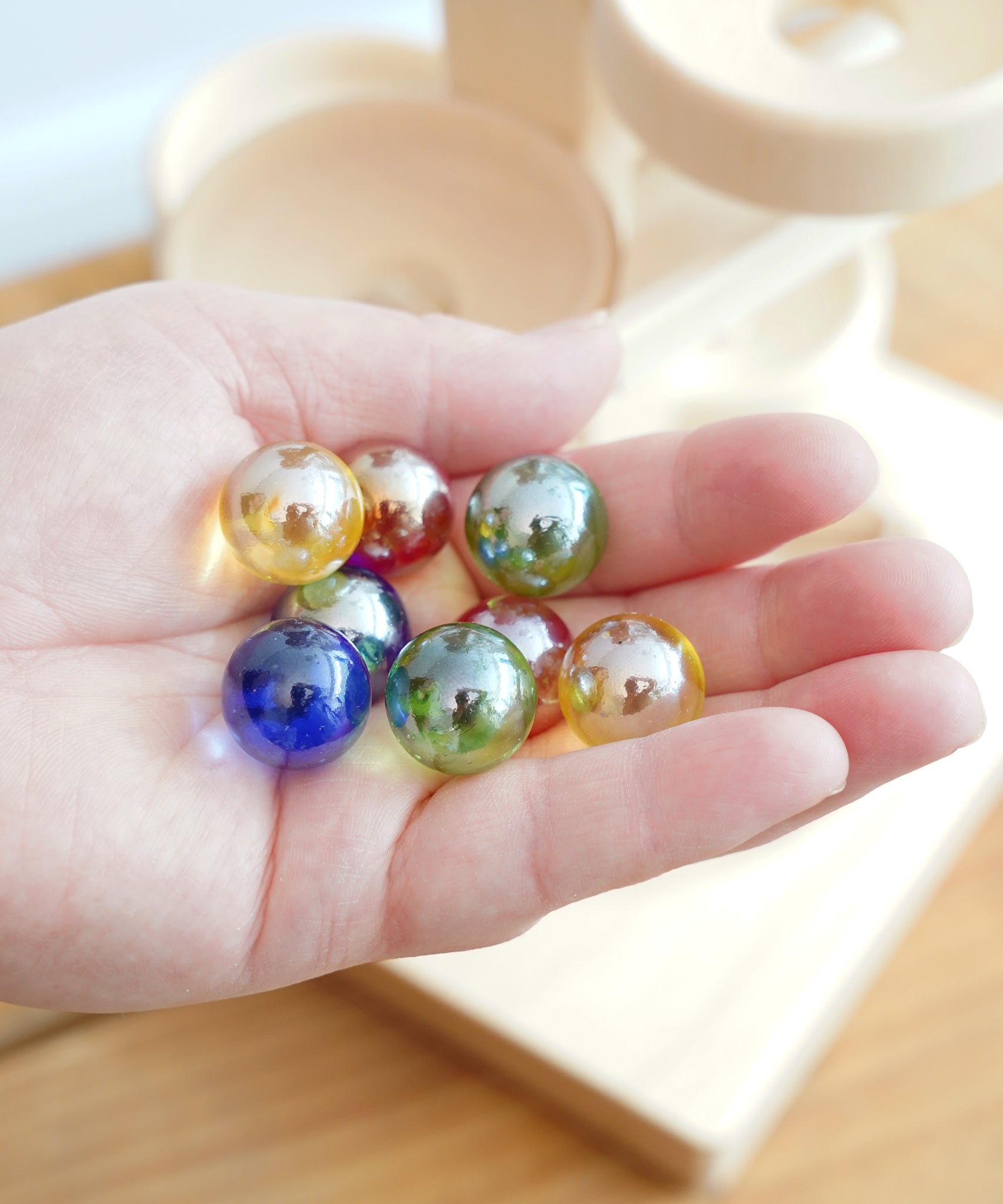 A close up of 8 coloured glass marbles from the Beck Toys Maple Wood Marble Run Plate Tower set in an adult's hand. 