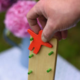 A close up of an adult's hand placing the red coloured wooden figure on the Beck Toy Wobbler wooden tumbling toy frame. 