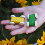 A close up of a yellow and a green coloured wooden race cars from Beck Toys in a child's hand. Sunflowers can be seen in the background.