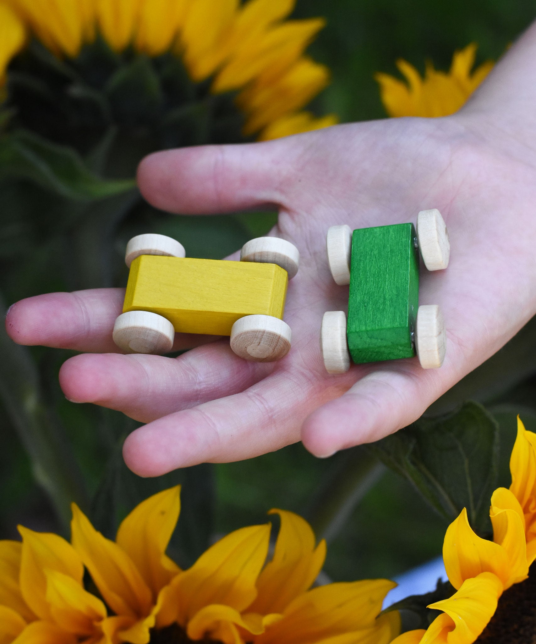A close up of a yellow and a green coloured wooden race cars from Beck Toys in a child's hand. Sunflowers can be seen in the background.