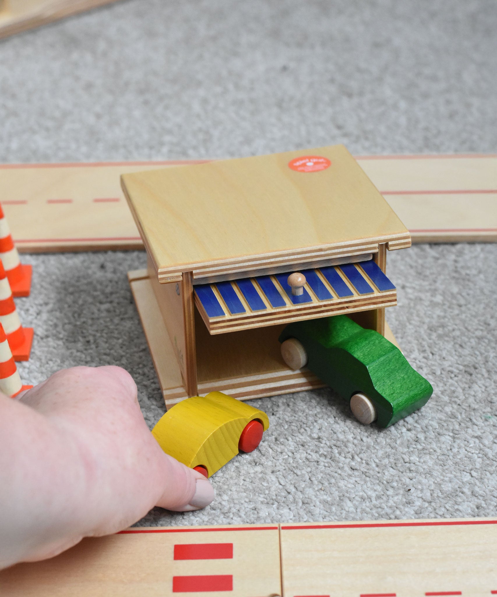 A Beck wooden toy car garage with a green coloured car half way in and a yellow one just outside. Beck have a range of wooden toys and ball runs available at Babipur. 