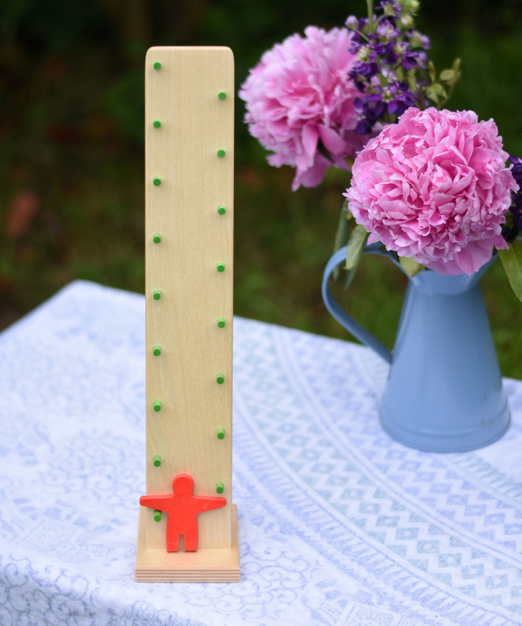 Beck wooden toys Wobbler tumbling toy with Red Figure placed on a small covered table. A bunch of flowers can be seen in the background. 