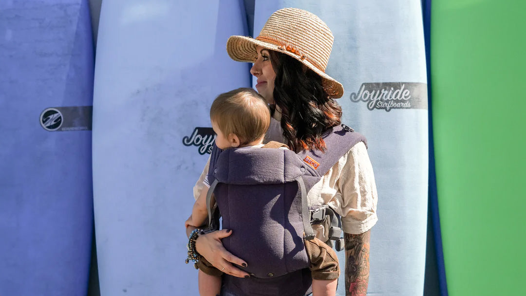 A mother carrying her baby in a Beco Carrier, on a summery day in front of some surfboards.