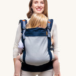 Beco Toddler Carrier - 20-60lb. The image shows an adult earing the carrier with a toddler inside. The carrier is navy blue with a mesh back, and orange detail, on a cream background