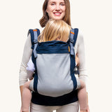 Beco Toddler Carrier - 20-60lb. The image shows an adult earing the carrier with a toddler inside. The carrier is navy blue with a mesh back, and orange detail, on a cream background