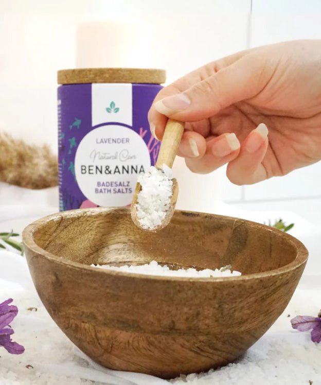 Ben & Anna lavender bath salts in a scooper being poured into a wooden bowl