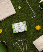 A bar of Bio-D dish washing soap inside it's green box lying on top of grass with various camping items. This dish soap bar is available at Babipur. 