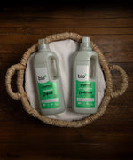 A 1L bottle of Bio-D fresh juniper fabric conditioner  and laundry liquid laying flat inside a washing basket. Both cleaning products available at Babipur. 