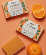 Bio-D mandarin soap bar lying next to mandarins on a light orange background. This soap bar is available at Babipur