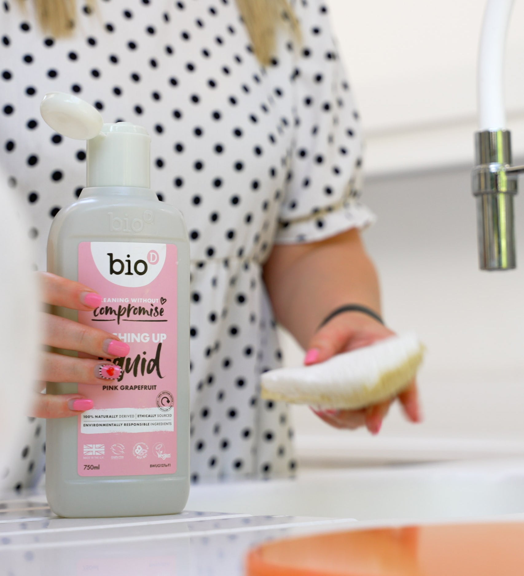 An adult holding the Bio-D pink grapefruit 750ml washing up liquid and washing the dishes. This washing up liquid is available at Babipur.
