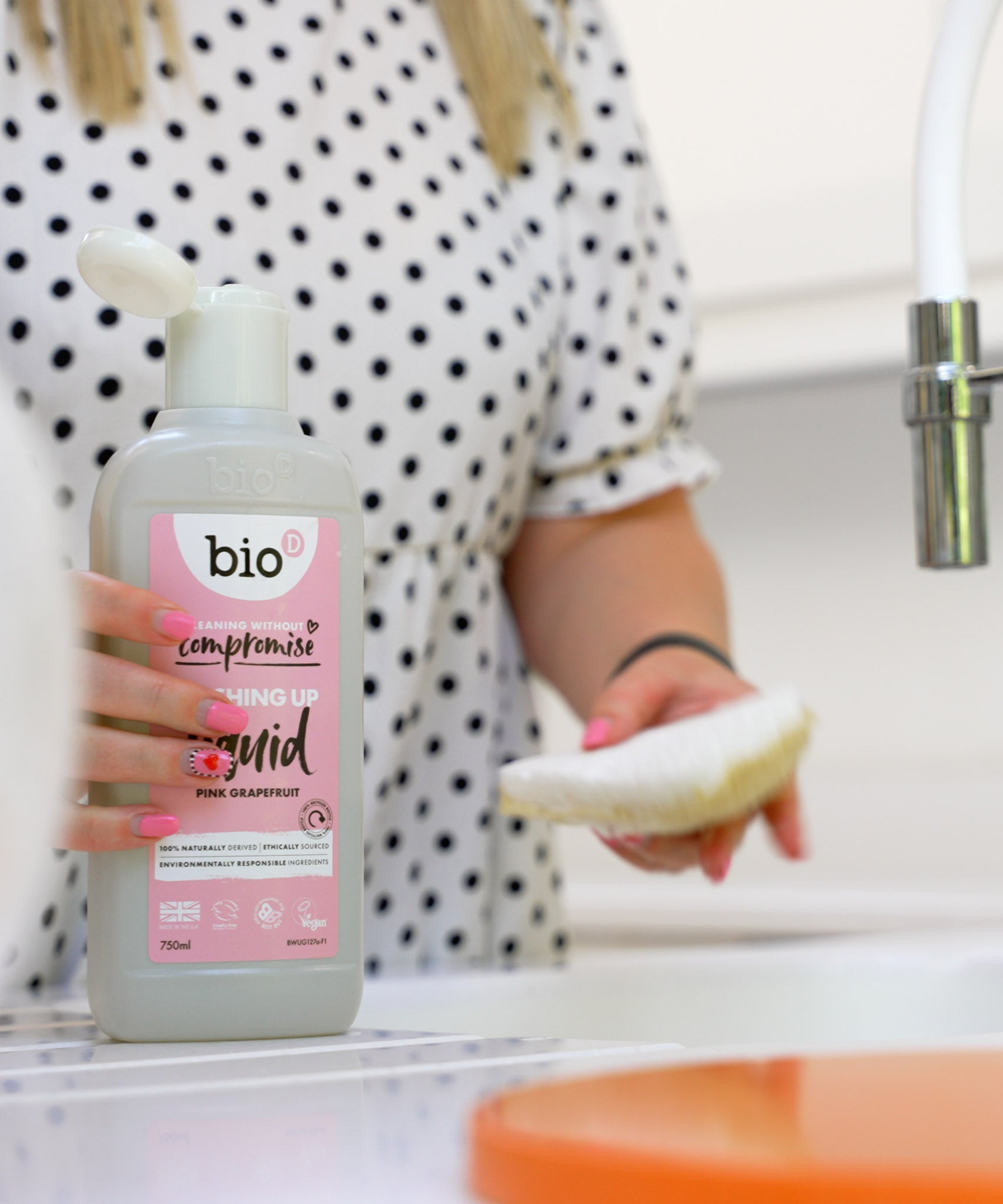 An adult holding the Bio-D pink grapefruit 750ml washing up liquid and washing the dishes. This washing up liquid is available at Babipur.