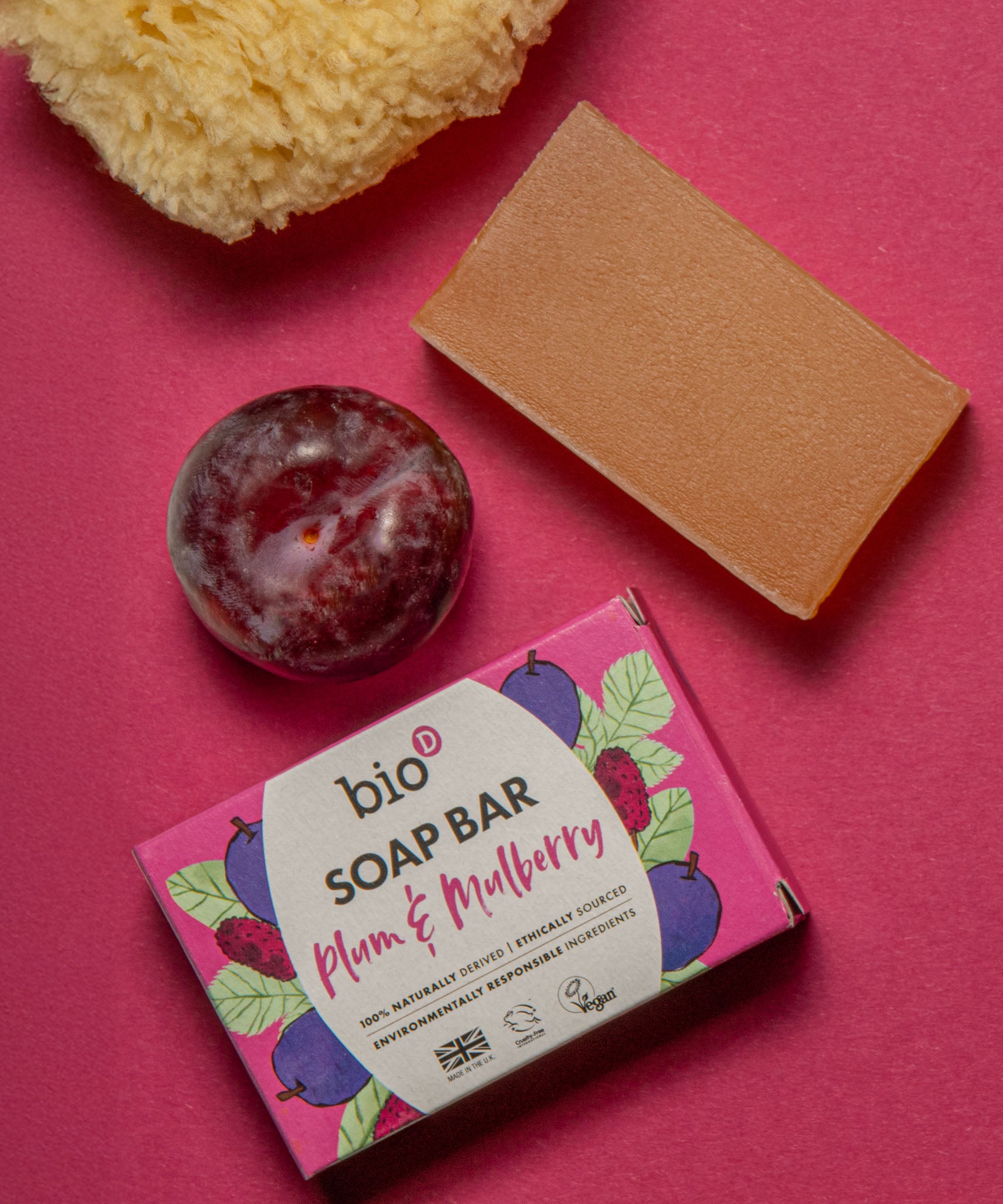 Bio-D plum & mulberry soap bar lying next to a plum and sponge on a light purple background. This soap bar is available at Babipur