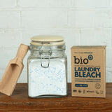 Glass storage jar filled with Bio-D Laundry Bleach powder beside the product’s cardboard box, with a wooden scoop shown to illustrate decanting and reuse.